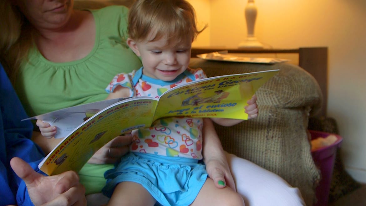 Toddler sits on her mother's lap and looks at a Curious George book.