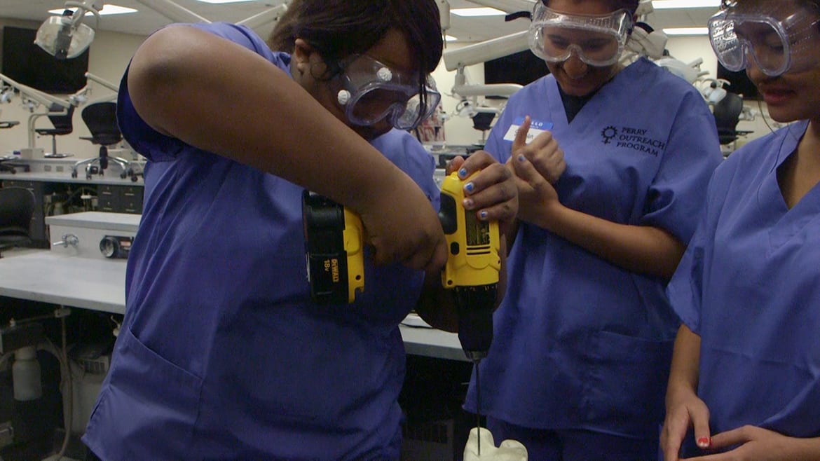 Three female high school students in scrubs and googles drill into an artificial bones.