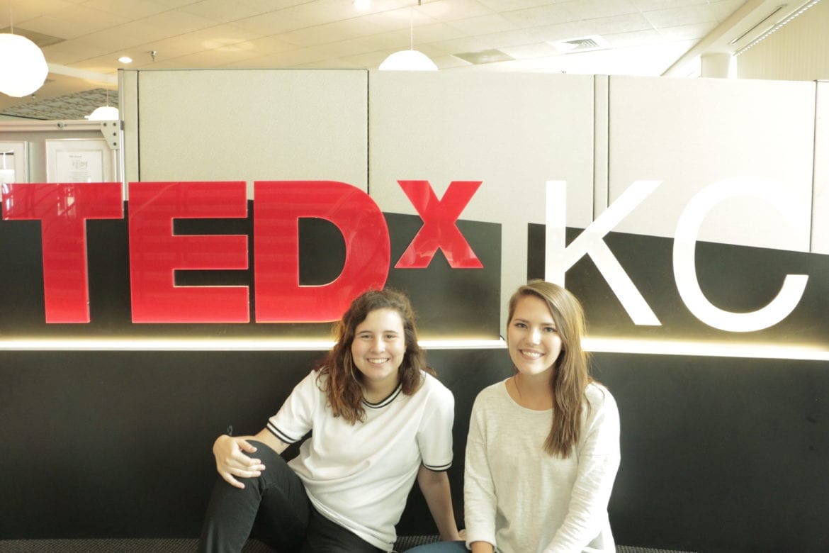 Two girls sit down in front of a TEDxKC sign indoors.