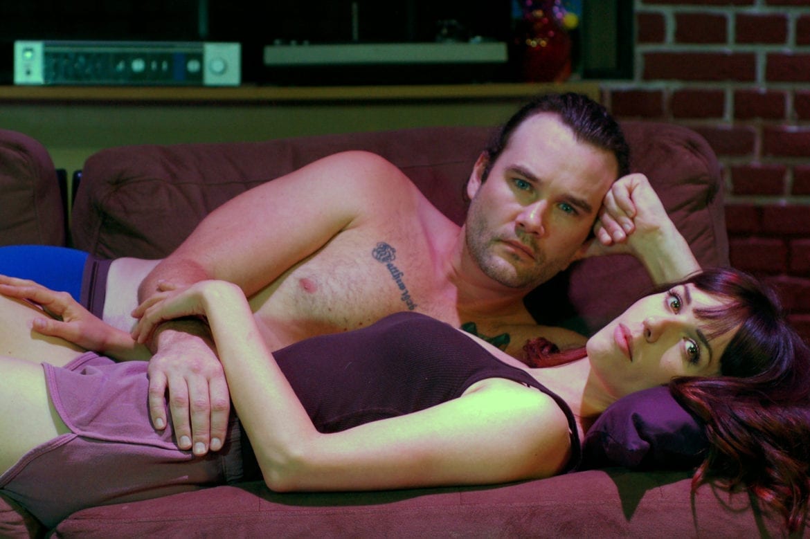 A man and woman laying on a sofa as part of a play, 