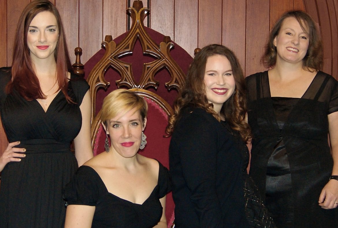 Four women sitting and standing in black dresses.