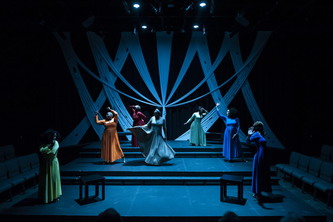 Seven women perform on a dim blue stage. 