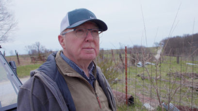 Man in a hat and glasses stands in front of rural l