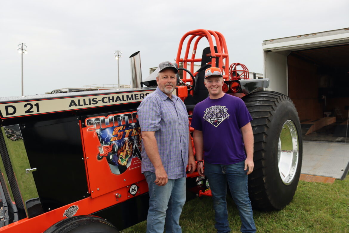 Two men in hats stand next to an orange tractor.