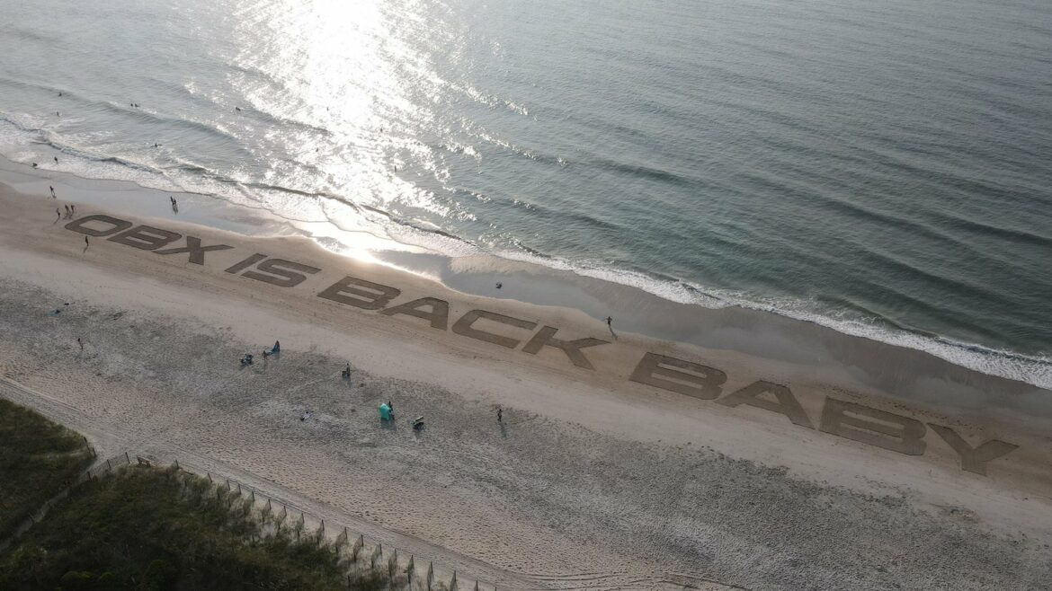 Letters carved into the sand on a beach read "OBX is back baby."