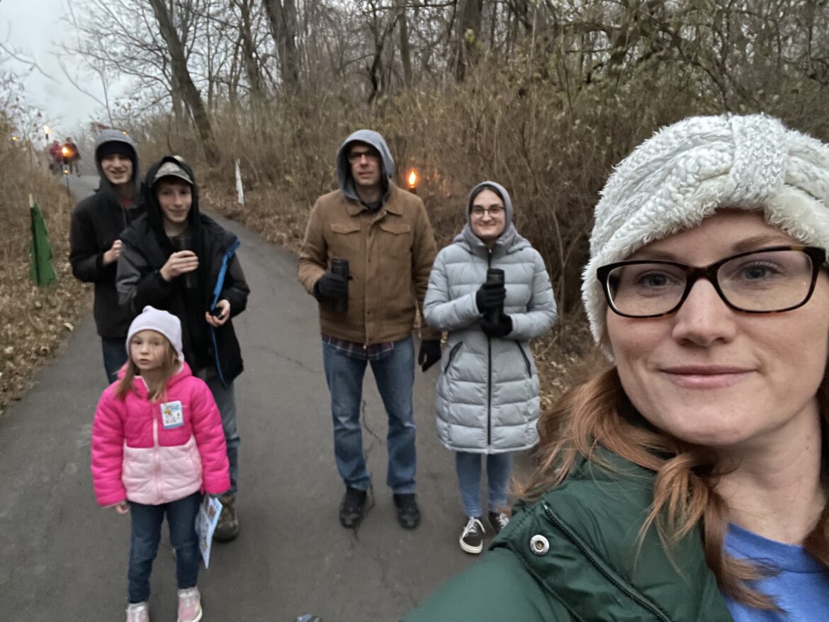 A woman with glasses takes a selfie on a nature trail, behind her are three kids and her husband. All are bundled up and holding hot chocolate.