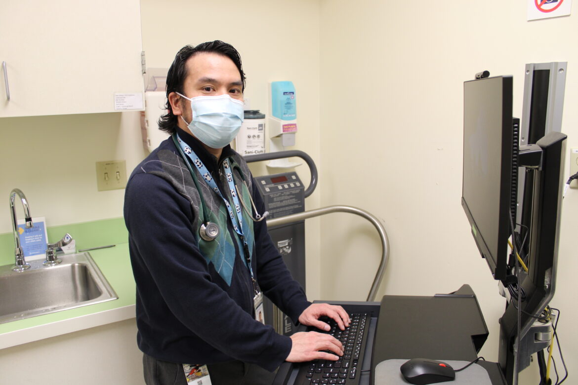 A young physician wearing a mask in his office, typing chart notes. Vibrant Health Center Dr. Carlos Garash Tamashiro serves a largely underserved, immigrant population.