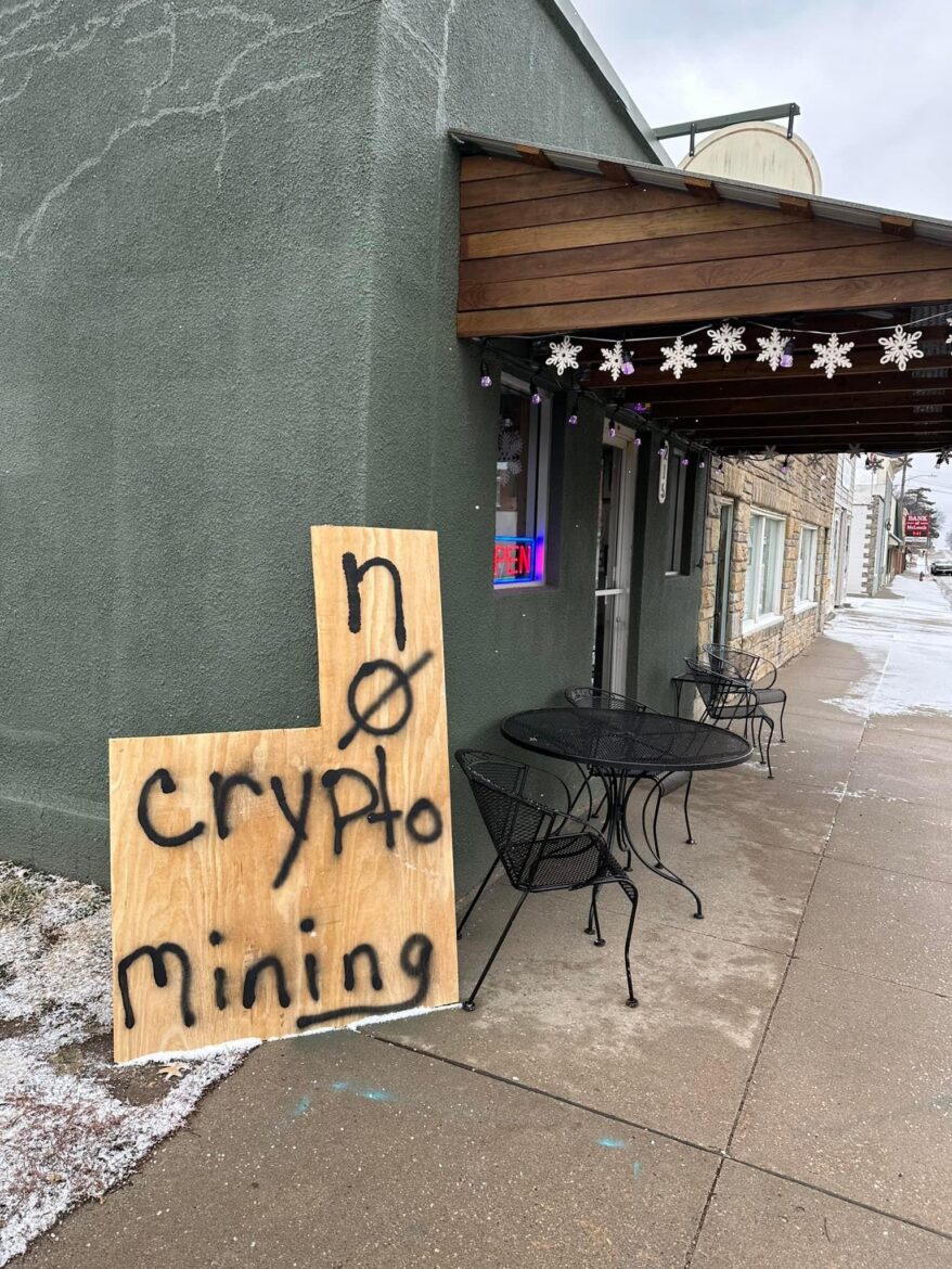 piece of junk leans against a building.  There are tables and chairs outside the restaurant and a "open" log in window.  The scrap wood is spray painted with "No crypto mining" 