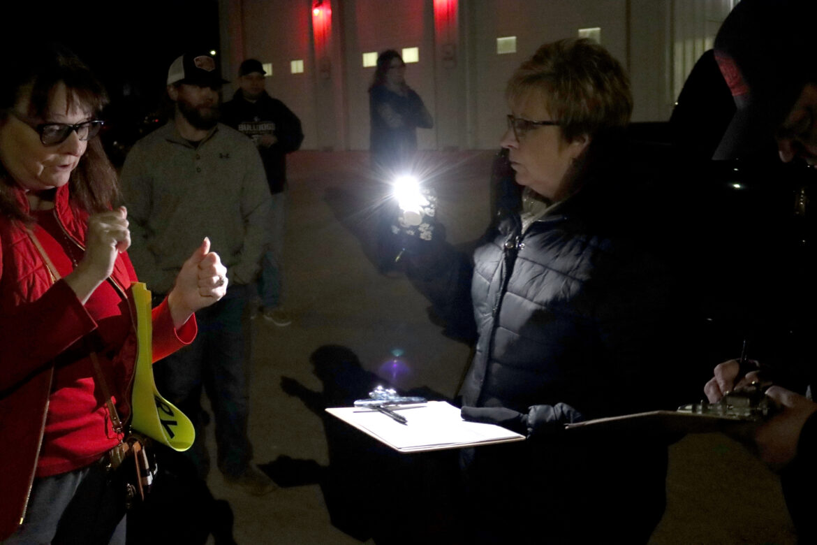 A woman in a black coat holds up a mobile phone with a flashlight on it.  Her other hand holds a clipboard and pen.  Next to her, someone is signing a petition and others are mingling around.