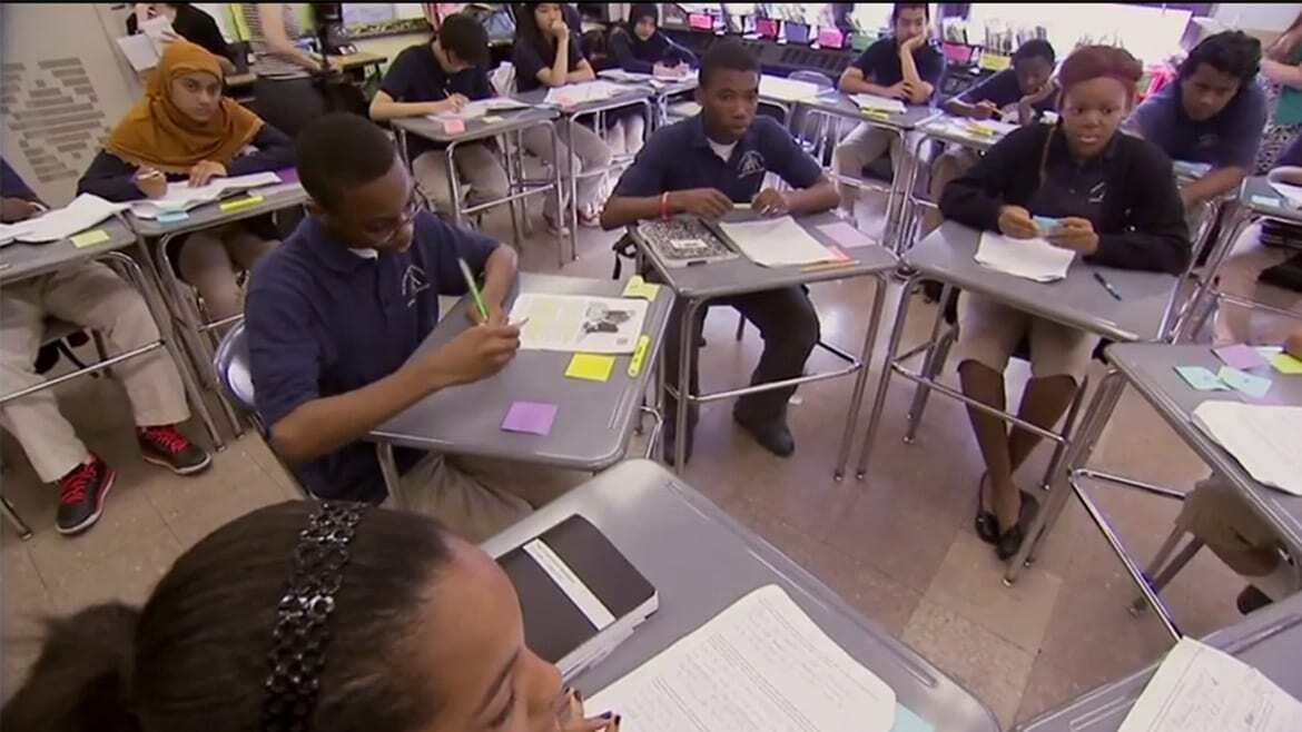 Students sit at desks in a circle and discuss assignment.