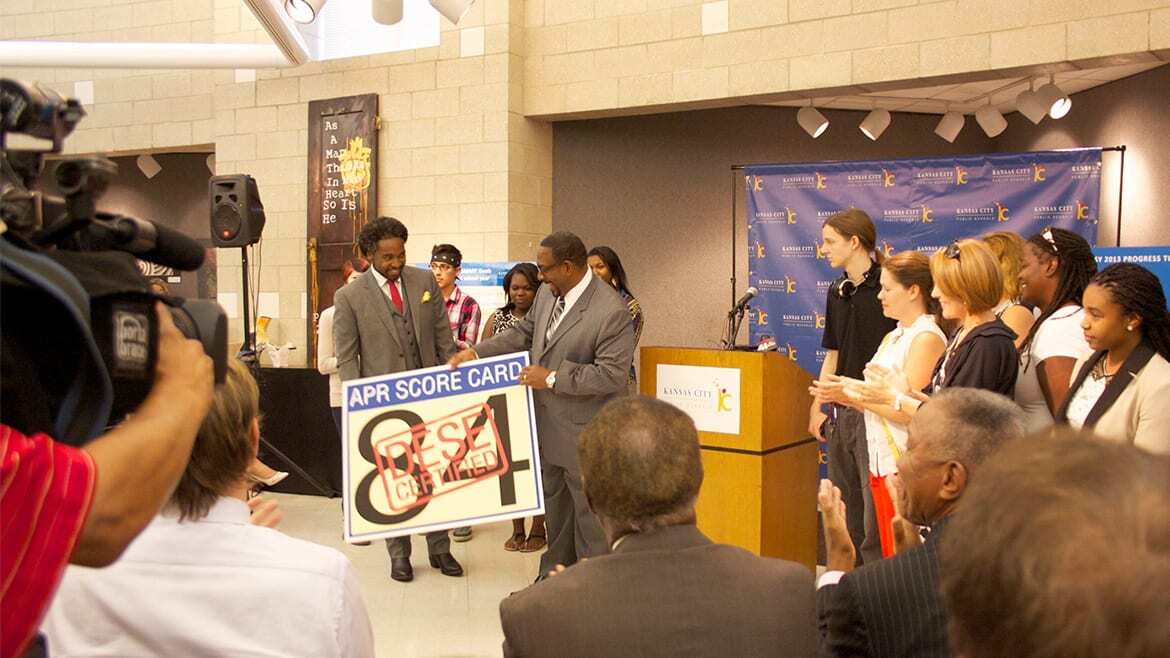 Camera and audience face podium where students watch as KCPS Superintendent Dr. Steve Green receives a large card with the district's 84 point APR score.