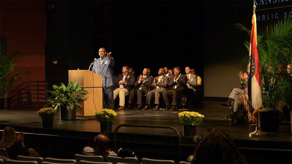Man stands at podium on stage with a hand outstretched to the audience.