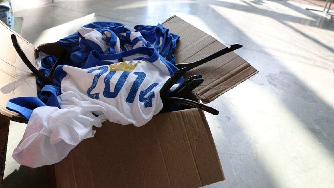 a pile of blue and white baseball T-shirts with "2014" (with a crown over the 0) overflow out of a box on a concrete floor.