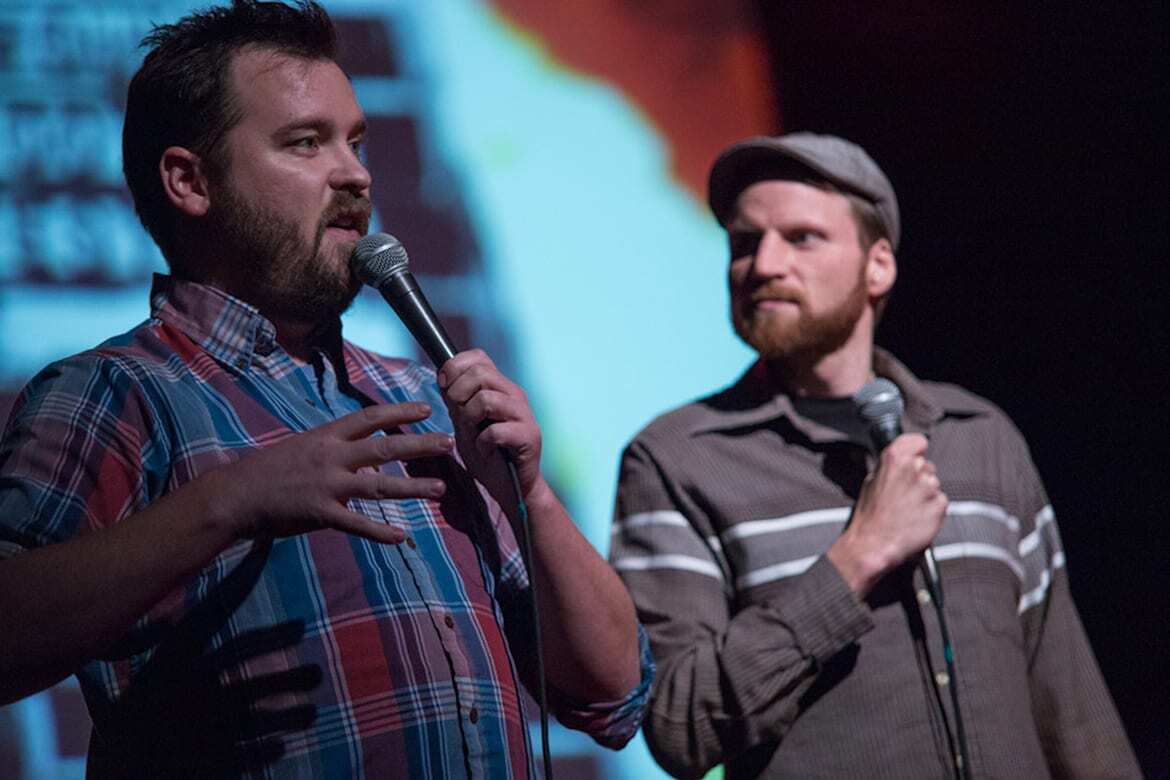 Picture of two men with microphones standing in front of a screen.