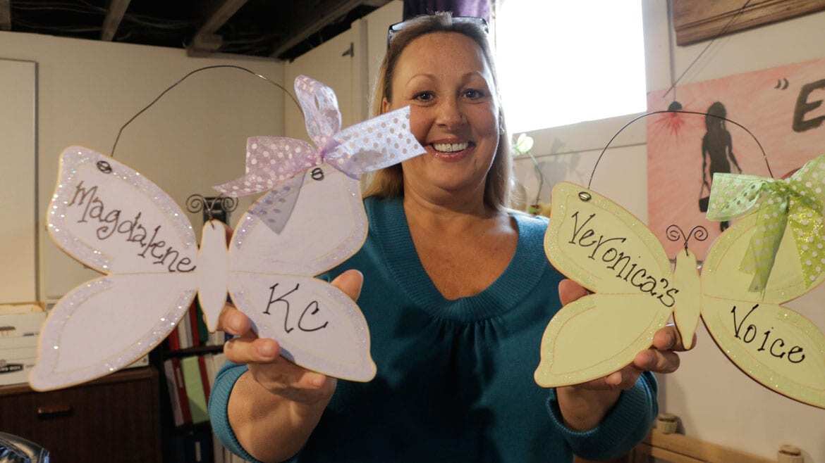 A woman in a blue sweater holds up two decorative butterflies. One says "Magdalene KC." The other says "Veronica's Voice."