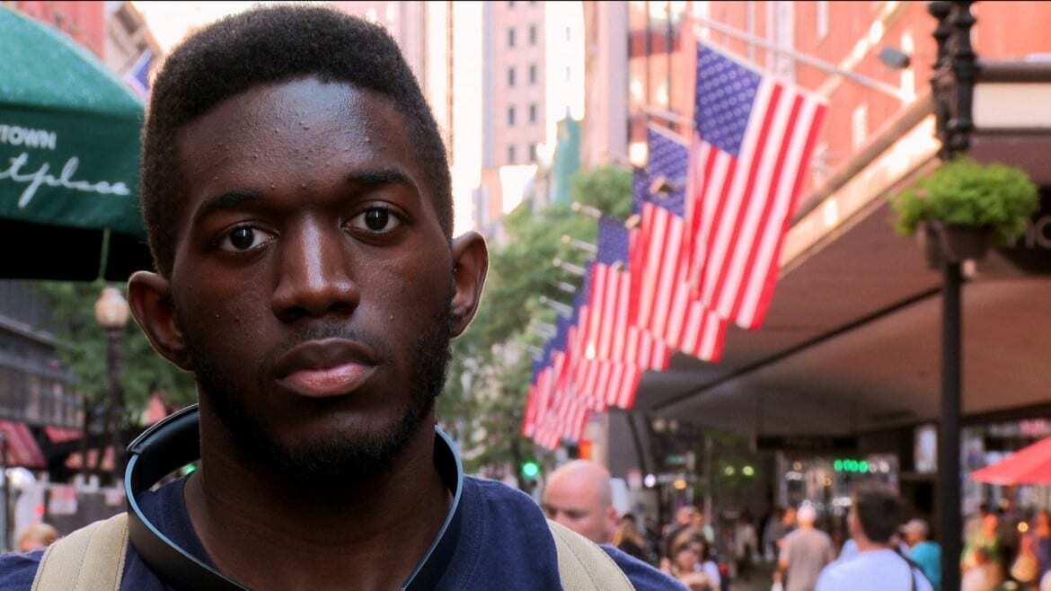 Image of African American man standing on street in front of row of American flags.