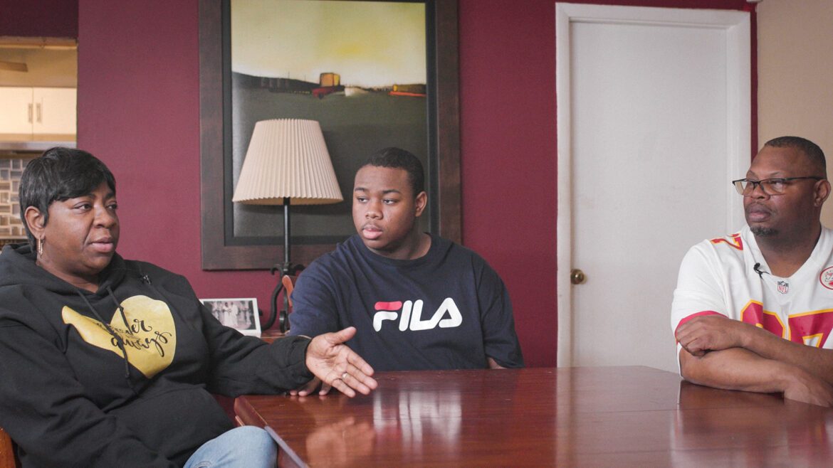 The Mitchell family sits around their kitchen table. Michelle is seated next to 17-year-old son with autism, explaining what she faces juggling work and finding support for her son.