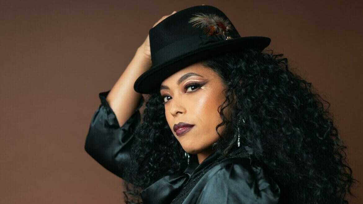 A woman with long curly hair, in a black silk shirt and black fedora hat looks into the camera.
