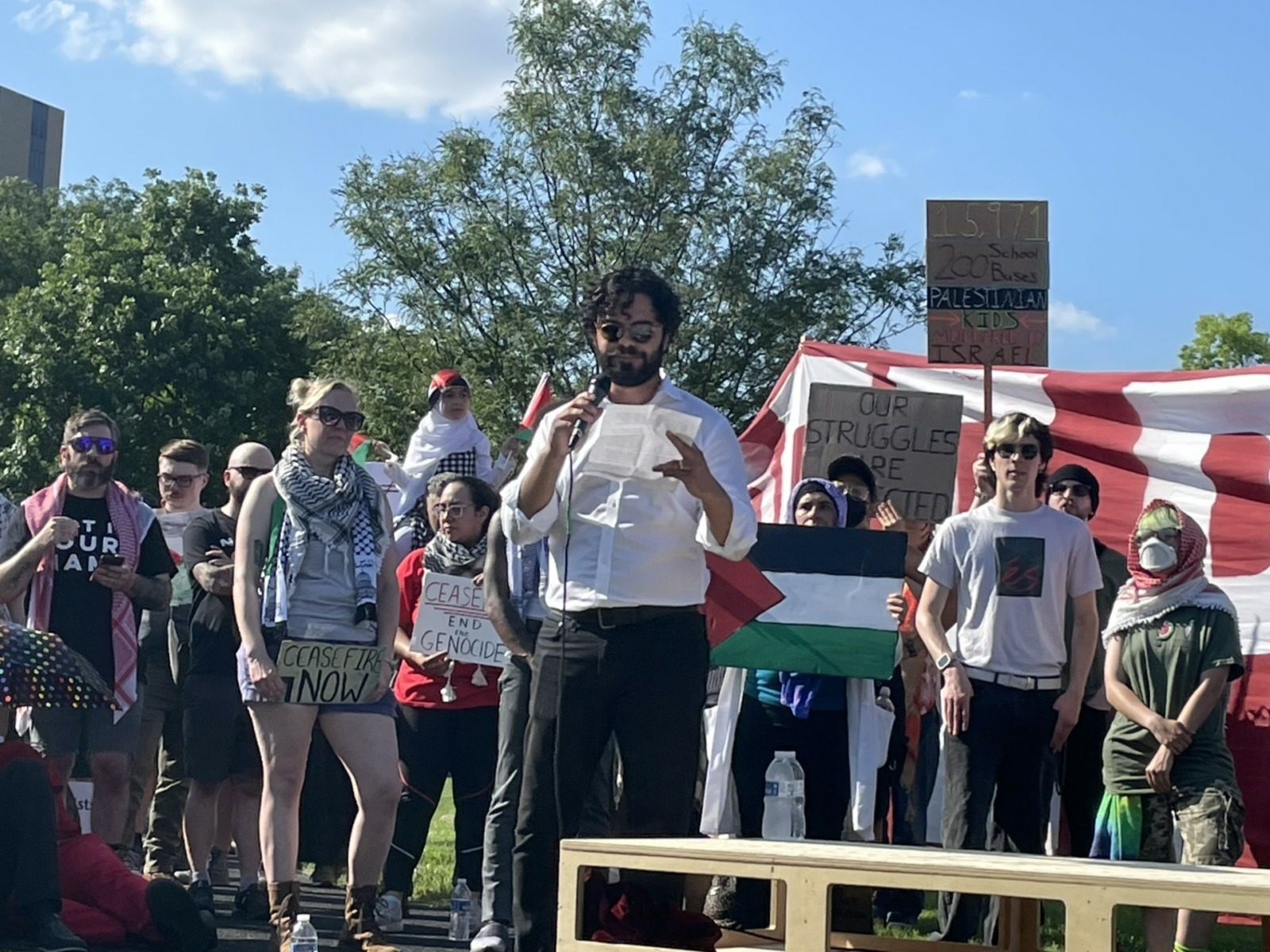 people standing outdoors. banners, palestinian flag, man reading from a white paper
