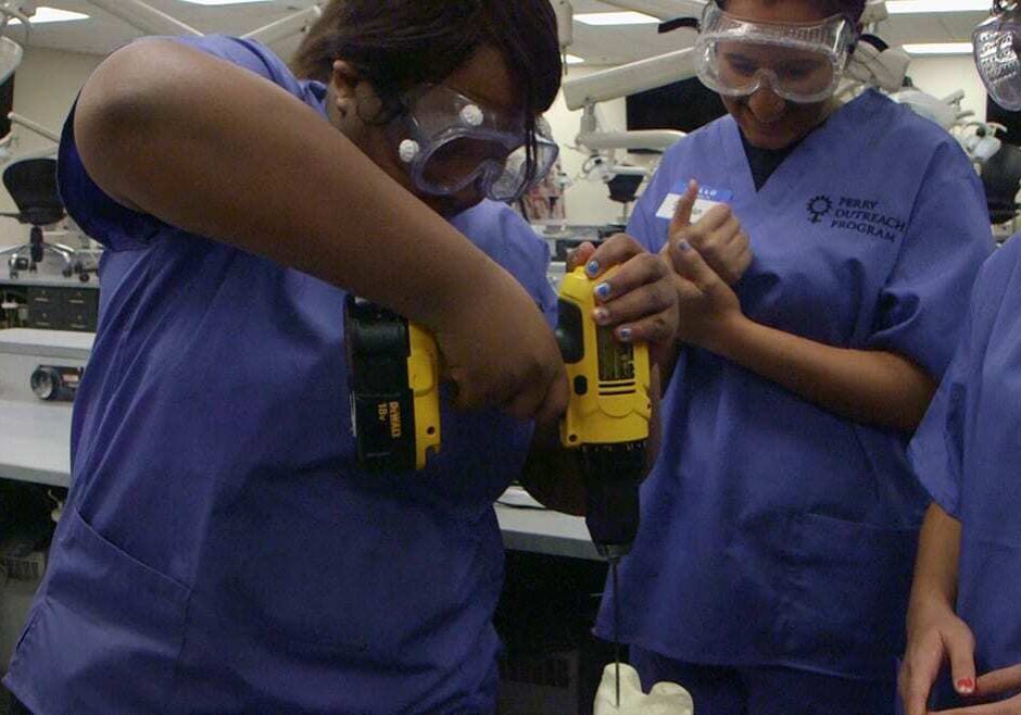 Three female high school students in scrubs and googles drill into an artificial bones.