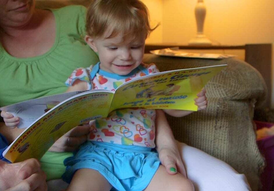 Toddler sits on her mother's lap and looks at a Curious George book.