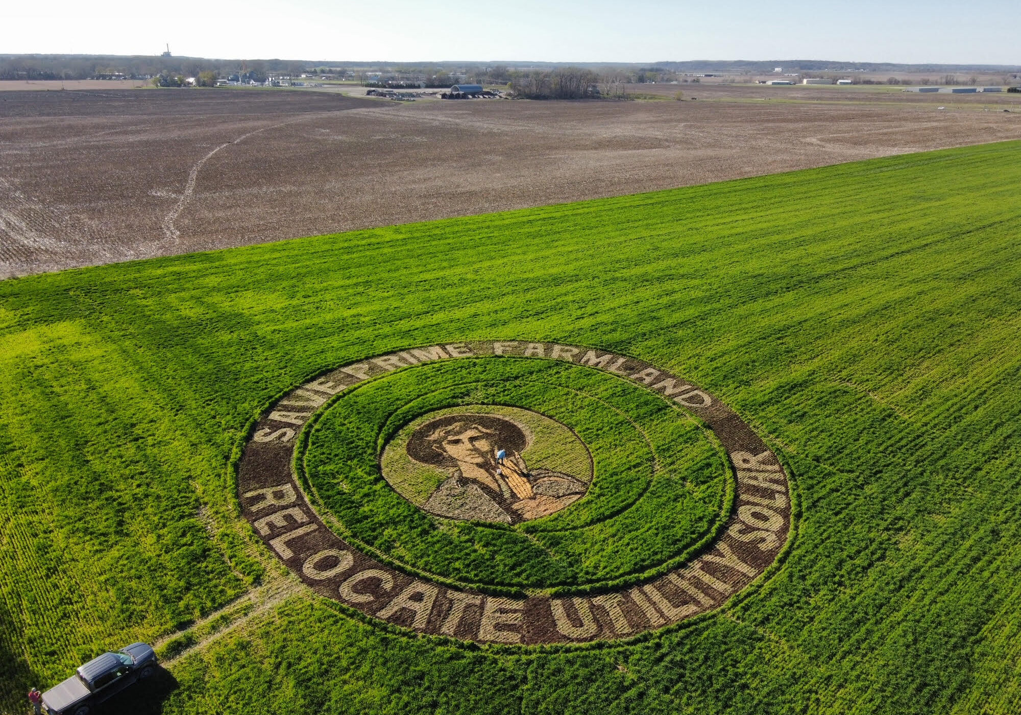 A green field. In the middle of the field is an art piece made of dirt and grass that depicts a young farmer. Around the outside of the circle it reads "Save Prime Soil" "Relocate Utility Soil"