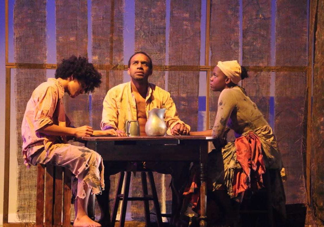 Three African American people sitting at a table depicting slaves. 