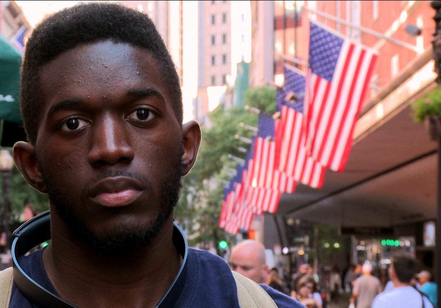 Image of African American man standing on street in front of row of American flags.