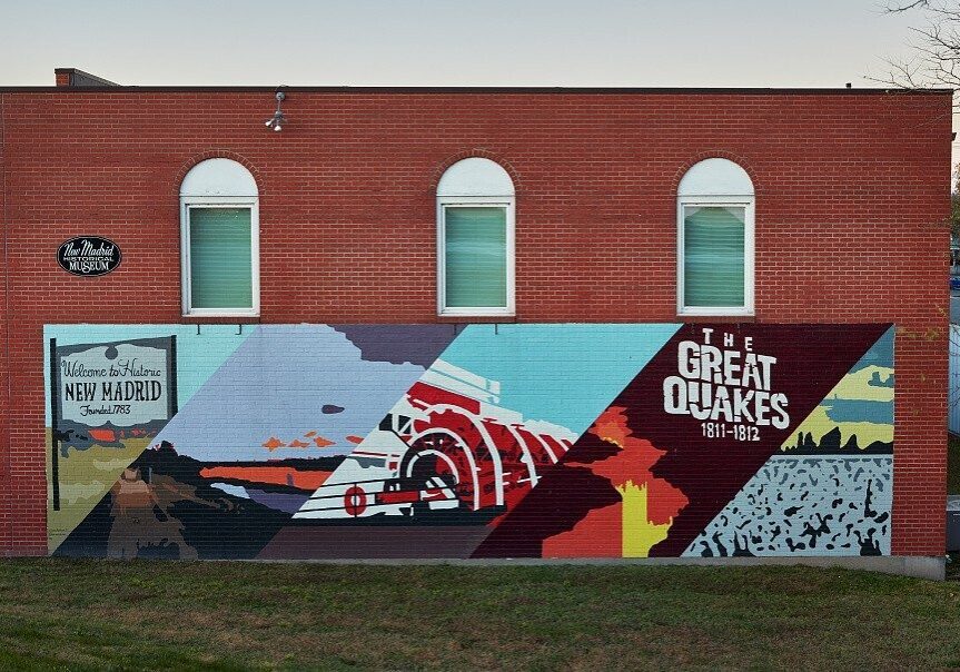 A colorful mural on the side of a brick building with the words "Welcome to Historic New Madrid" and "The Great Quakes" at dusk